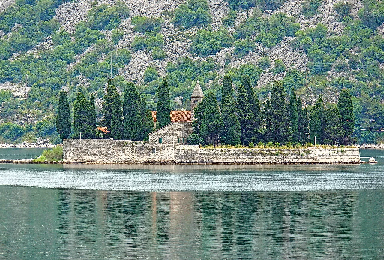 The islet of St George near Perast, with cypresses, a church and a stone wall on calm green water