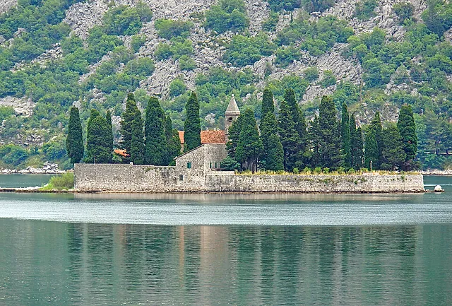 The islet of St George near Perast, with cypresses, a church and a stone wall on calm green water