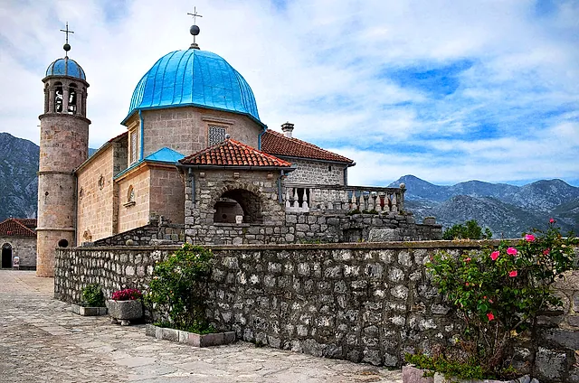 The blue-domed church of Our Lady of the Rocks on its islet, seen over a stone wall with roses