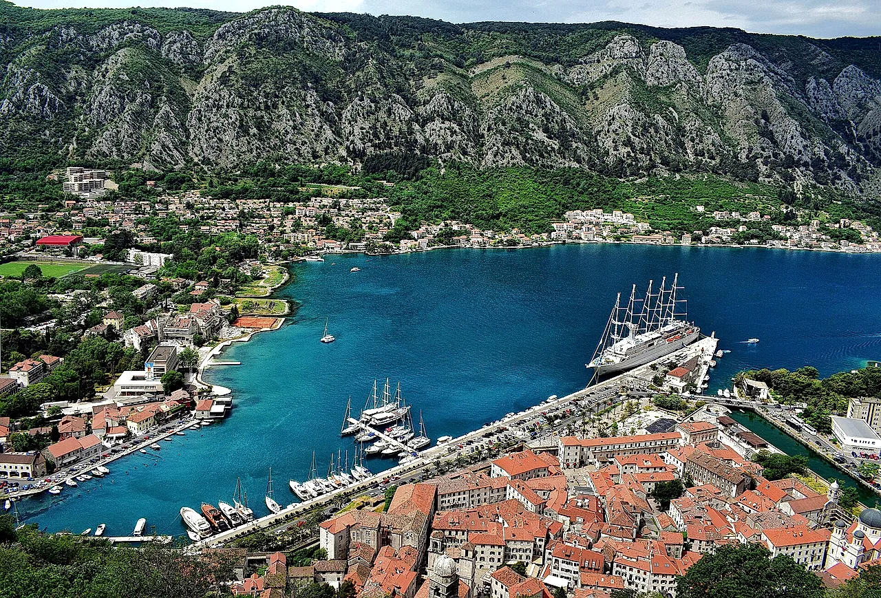 Kotor’s red-roofed old town beside the blue bay, with a tall ship moored and mountains behind